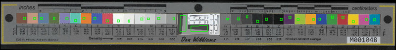 Gray rectangular bar oriented horizontally with gray slanted rectangles outlined in green near the center, dots on the edges, and squares of colors and white, gray and black values aligned from left to right that have corresponding numerical values printed under them (LAB colorspace values) with little green outlined squares within them, the green squares a product of the target being measured for FADGI compliance by software.