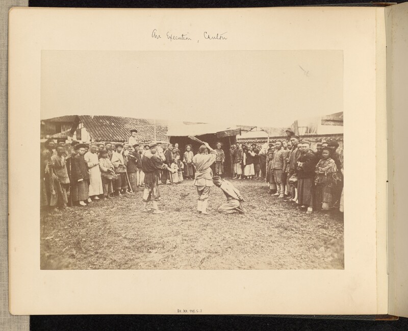 A photograph of a crowd circled around a kneeling man with two men pointing weapons at him.
