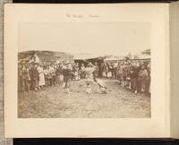 A photograph of a crowd circled around a kneeling man with two men pointing weapons at him.