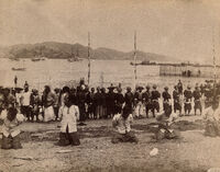 A black and white photograph of 5 men on a beach kneeling with their hands behind their back with a large crowd behind them.